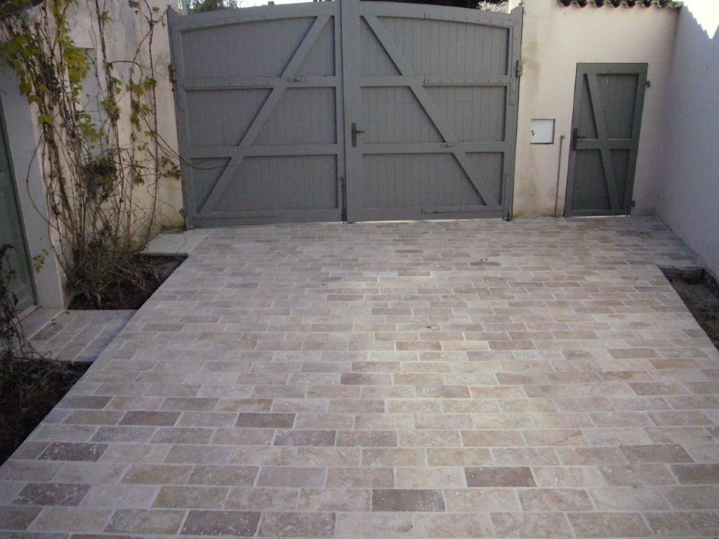 Allée pavée menant à un portail métallique gris fermé, situé à côté d'un mur blanc et d'une porte latérale.