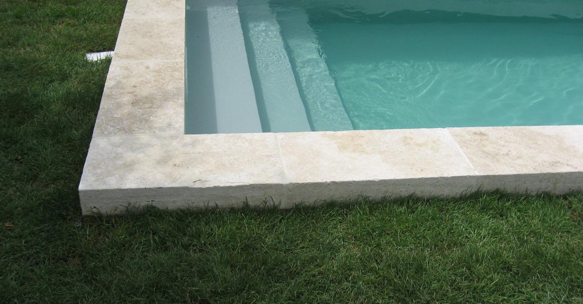 Piscine bordée de pierres, jouxtant une pelouse verdoyante, aux eaux turquoise limpides et dotée d'un escalier d'angle.