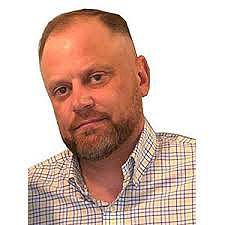 Mann mit kurzem Haar und Bart, der ein kariertes Hemd trägt, schaut in die Kamera.