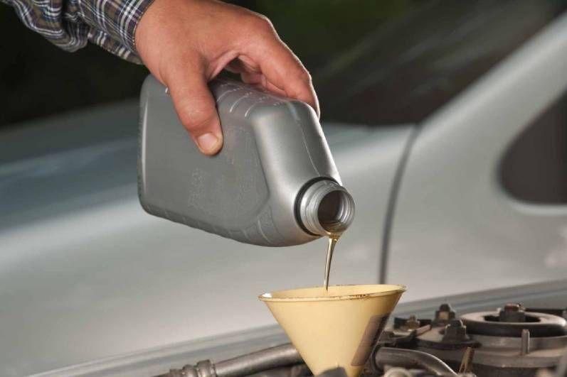 Une main verse de l'huile moteur dorée d'une bouteille en plastique grise dans un entonnoir jaune placé dans le compartiment moteur d'une voiture.