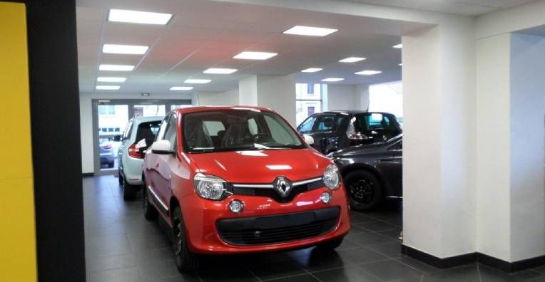 Une Renault Twingo rouge vif est garée dans le hall d'exposition bien éclairé d'une concession automobile, d'autres véhicules étant visibles à proximité.