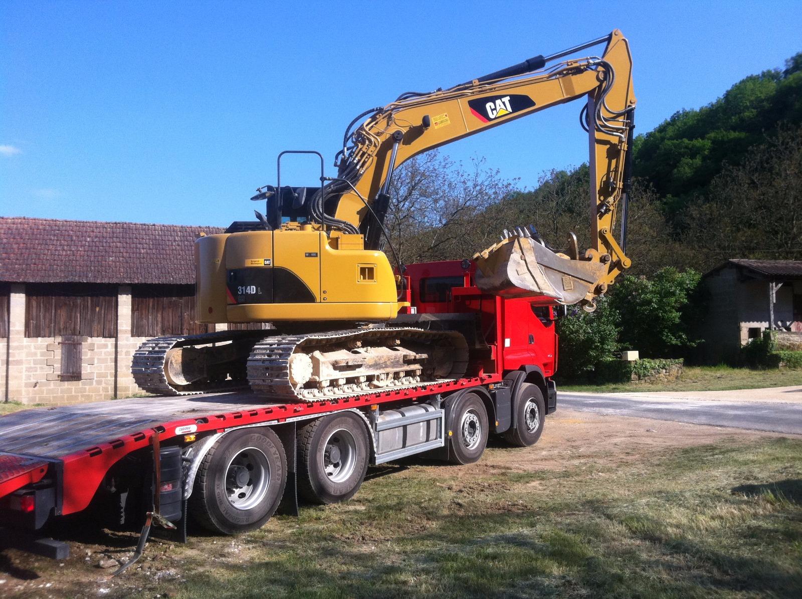 Une pelle mécanique jaune est chargée sur un camion plateau rouge dans une cour, avec un bâtiment et des arbres en arrière-plan.