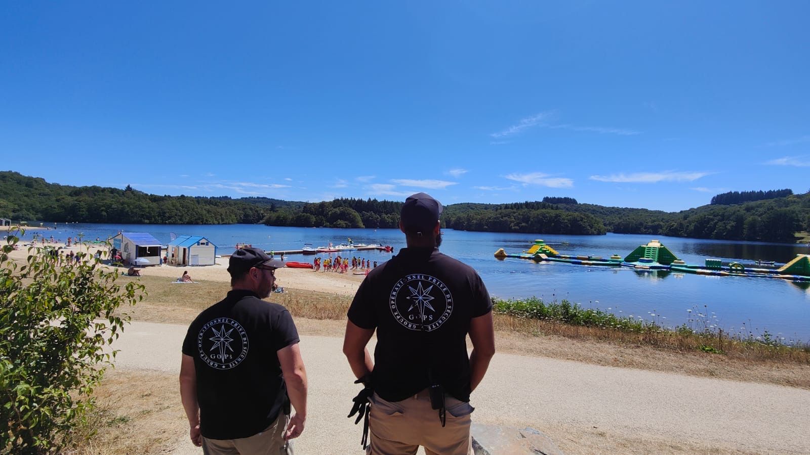 Deux hommes portant des chemises noires avec un logo se tiennent sur un chemin surplombant un lac avec des structures gonflables flottantes par une journée ensoleillée.