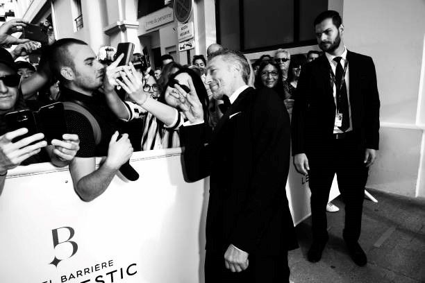 Un homme bien habillé salue les fans et prend des selfies. Un agent de sécurité les observe alors qu'ils se tiennent devant un bâtiment.