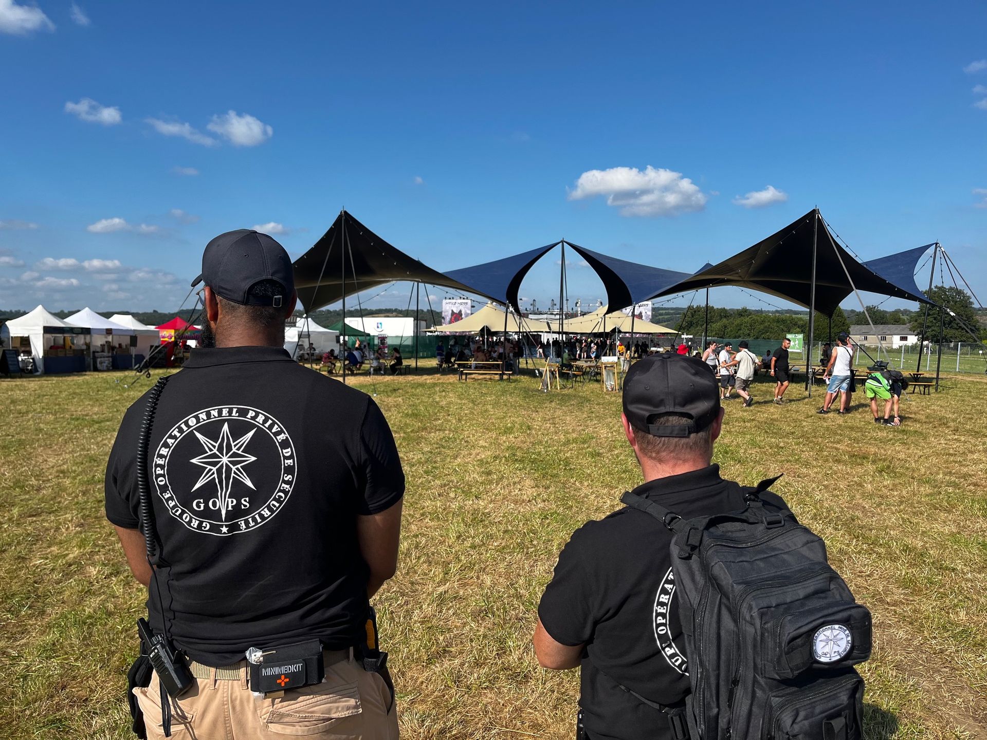 Deux agents de sécurité surveillent la foule lors d'un événement en plein air. Ciel bleu, tentes noires et champ herbeux.