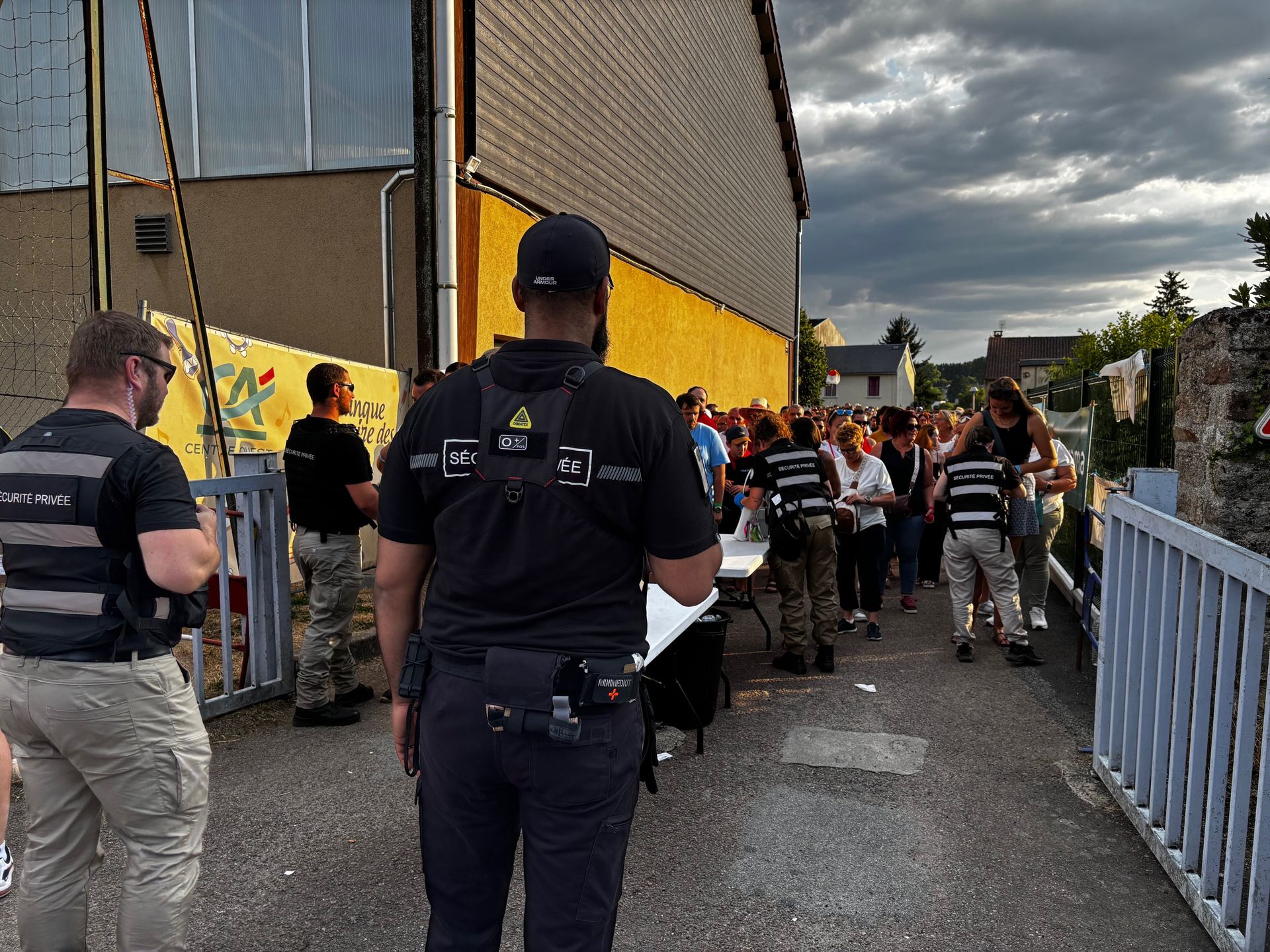 Des agents de sécurité près d'une foule, vérifiant probablement les pièces d'identité, sous un ciel nuageux.