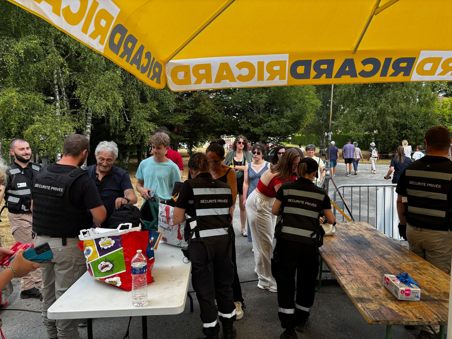 Des personnes se rassemblent lors d'un événement en plein air sous un parasol jaune Ricard. Le personnel de sécurité assiste les participants à une table.