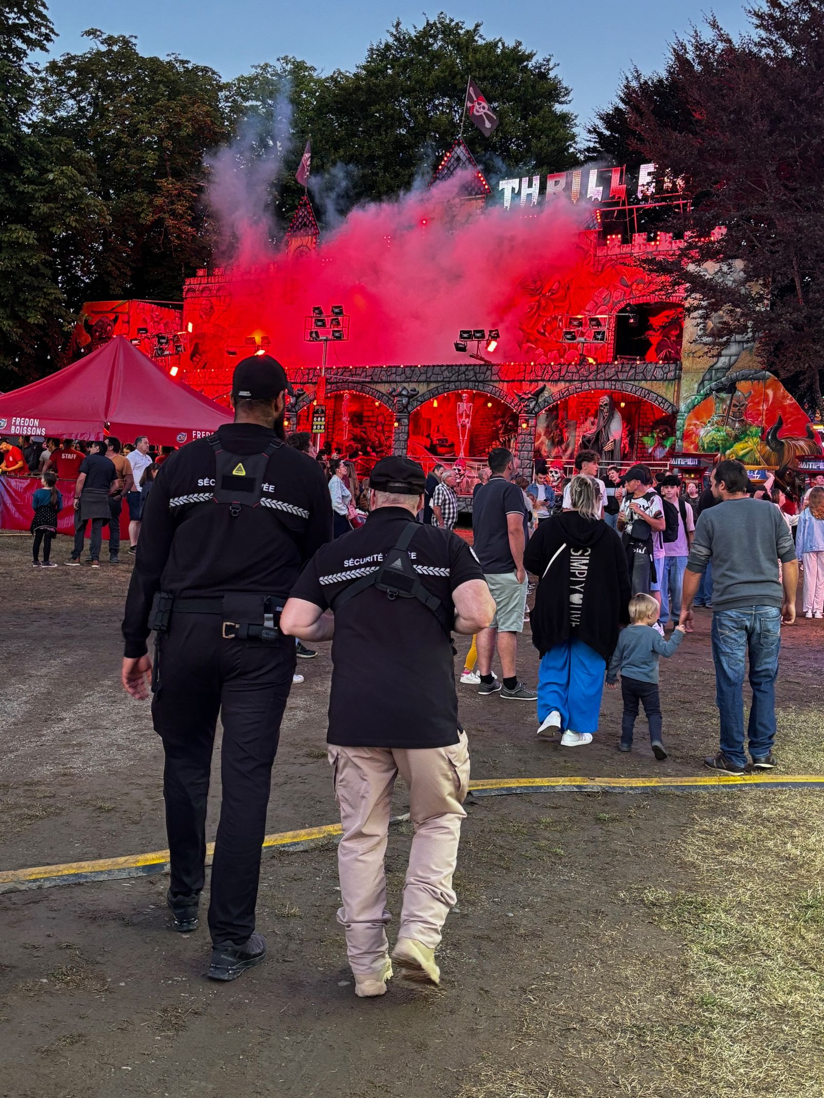 Deux agents de sécurité escortent une personne vers une scène avec de la fumée rouge et une foule lors d'un événement en plein air.
