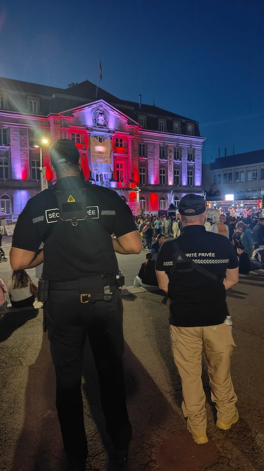 Deux agents de sécurité montent la garde sur une place publique au crépuscule ; le bâtiment est éclairé en rouge et violet en arrière-plan.