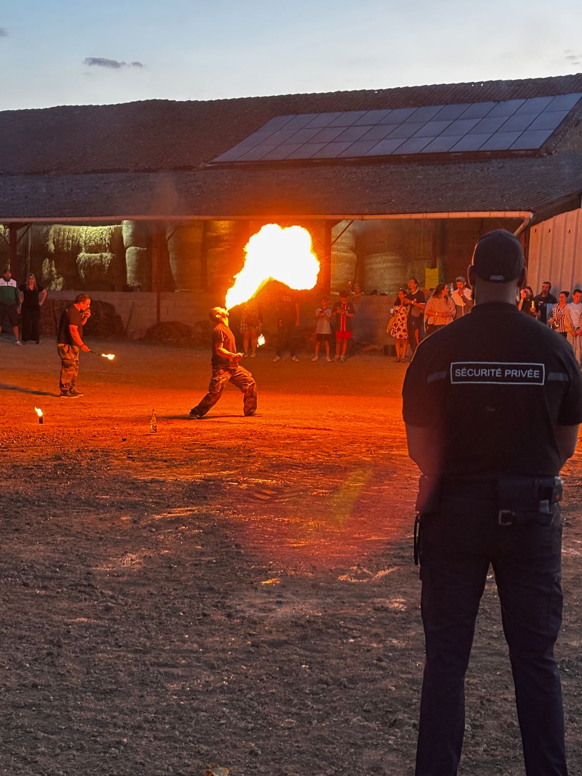 Des artistes du feu crachant du feu lors d'un événement en plein air, sous le regard d'un agent de sécurité.