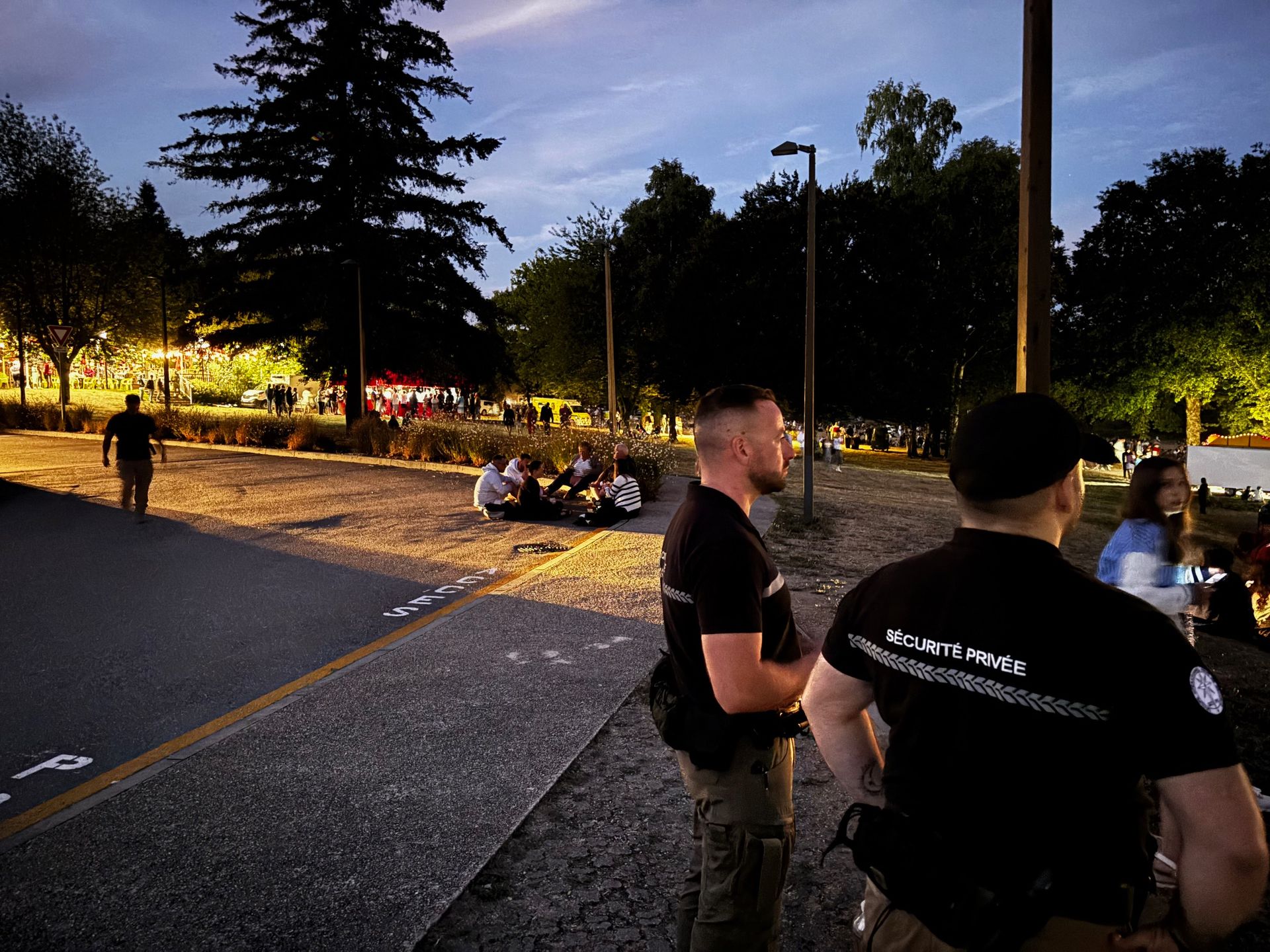 Deux agents de sécurité montent la garde près d'un groupe de personnes assises sur un chemin dans un parc au crépuscule.