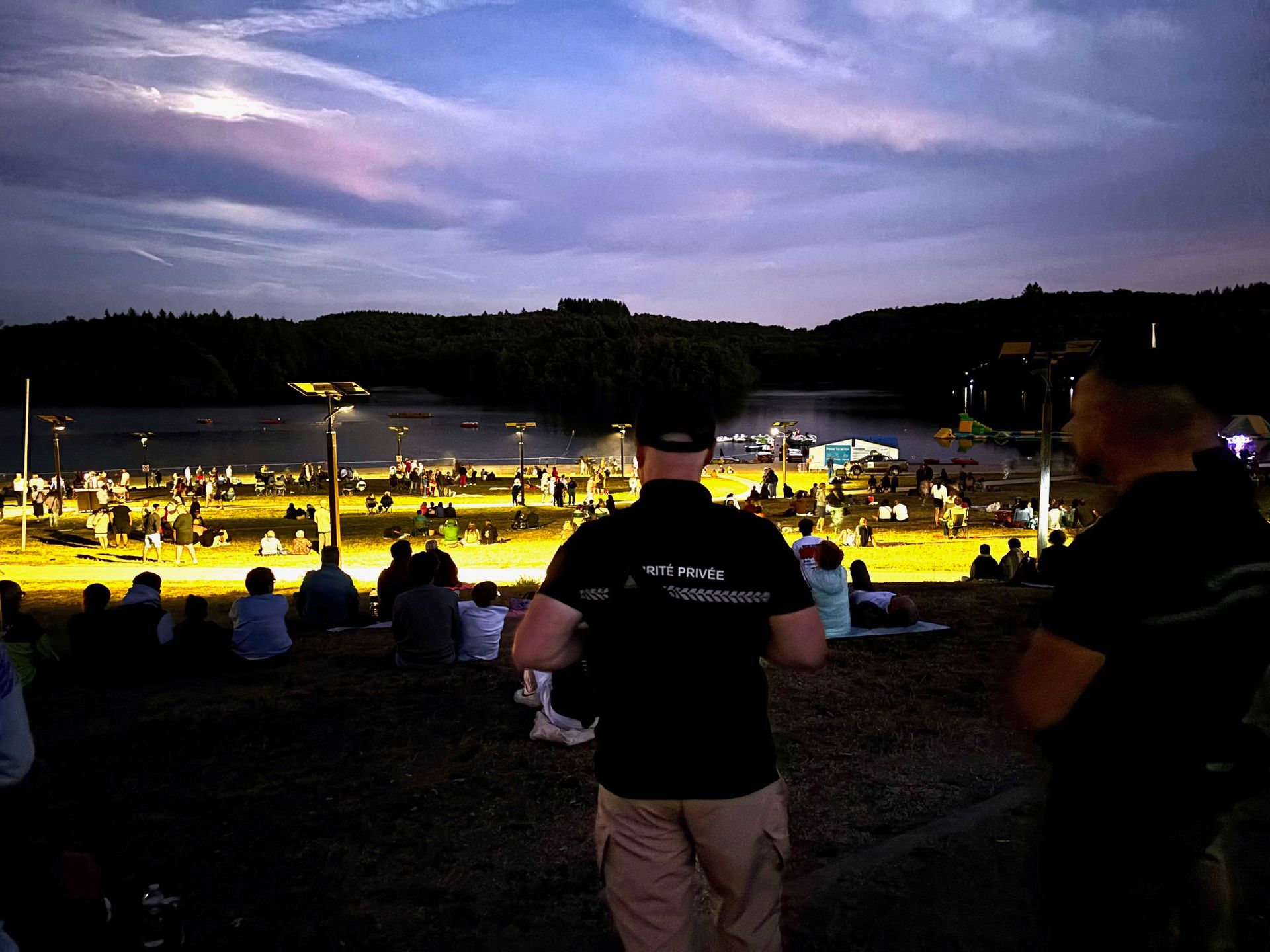 Des gens regardent un événement au crépuscule, une grande scène au loin, des silhouettes sombres au premier plan, un ciel violet, un décor en plein air.