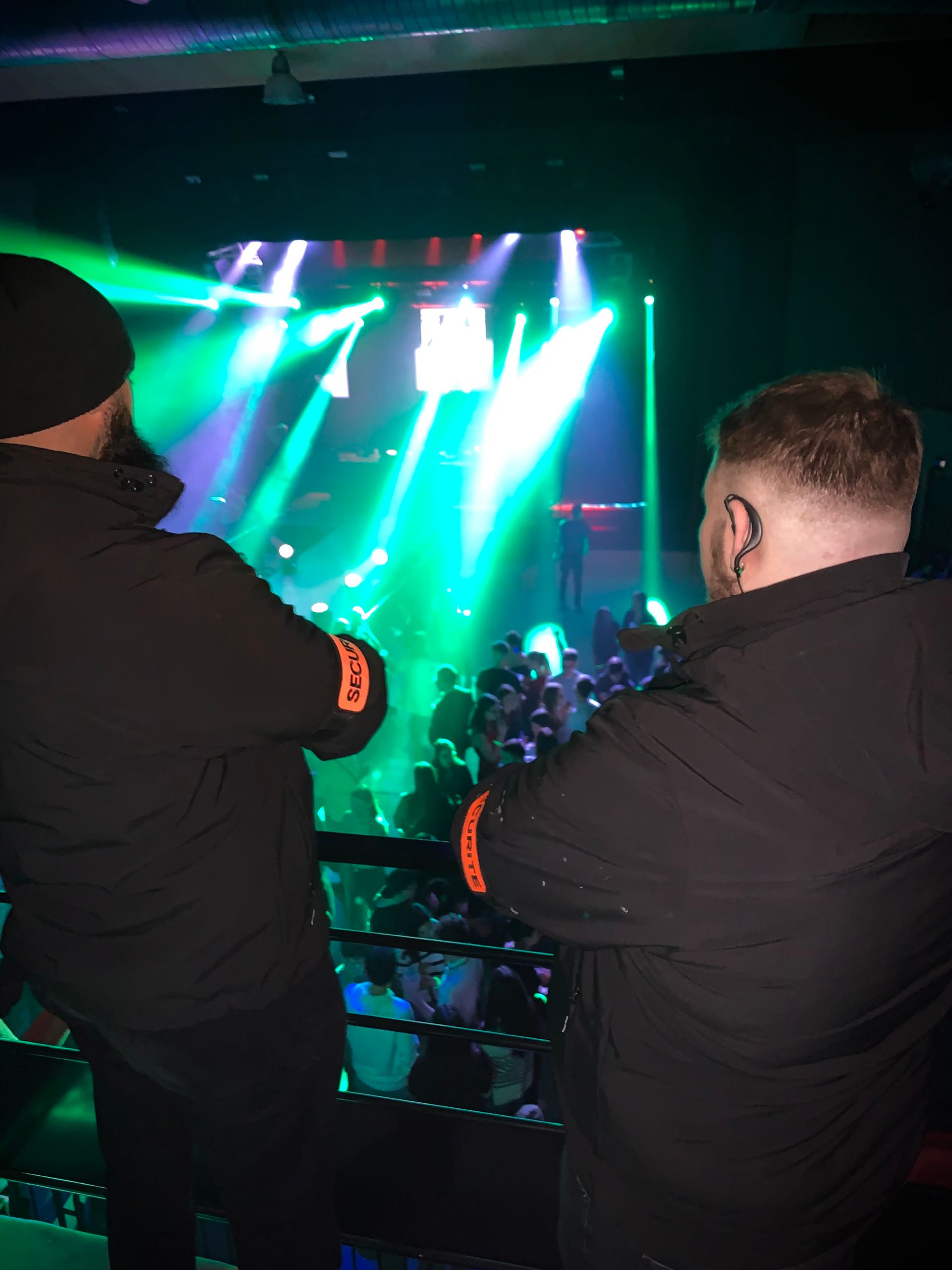Deux agents de sécurité surveillent la foule sur la scène d'une boîte de nuit avec des lumières vertes et bleues.