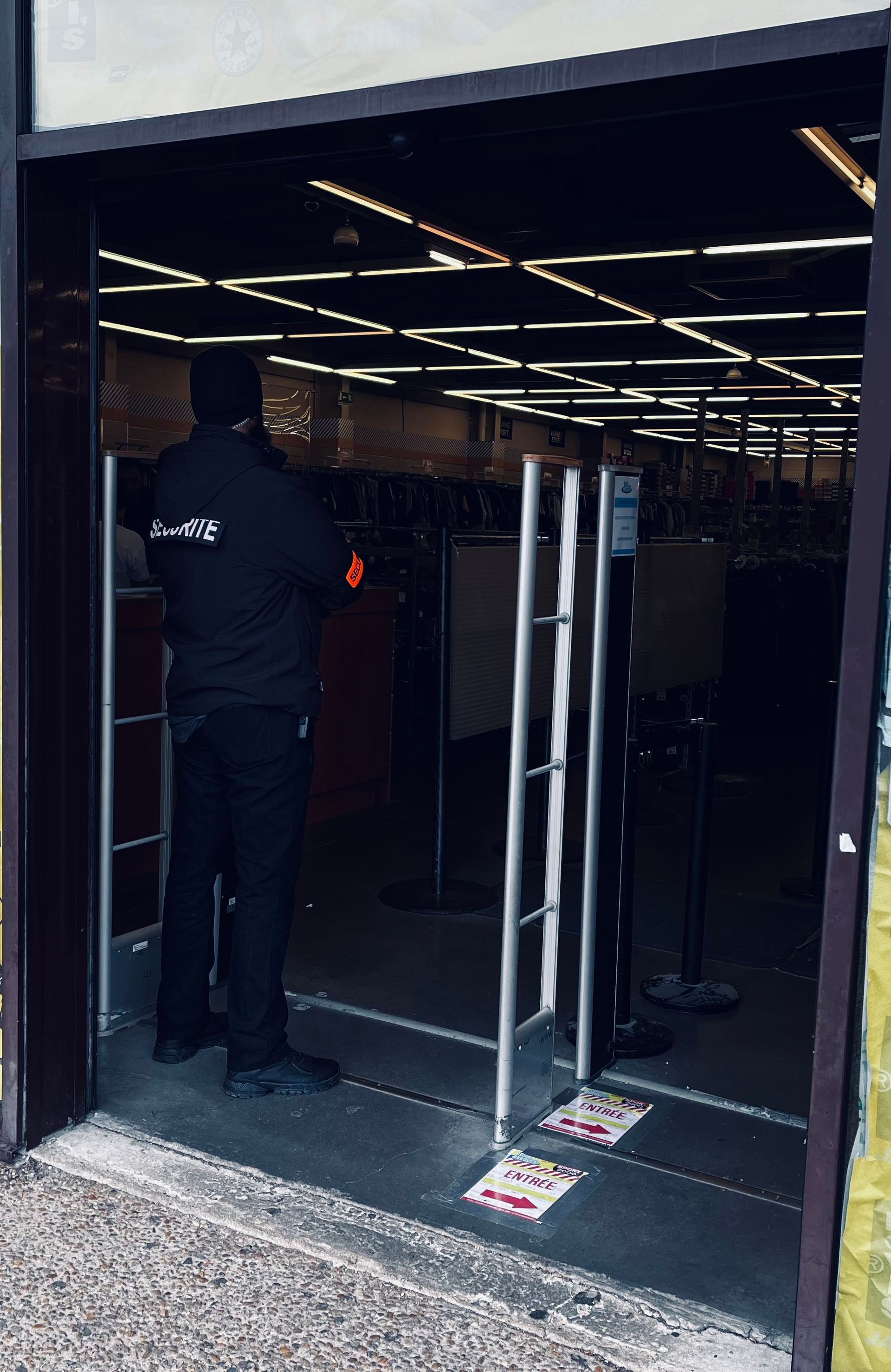Un agent de sécurité en uniforme noir se tient à l'intérieur de l'entrée d'un magasin, près des barrières antivol.