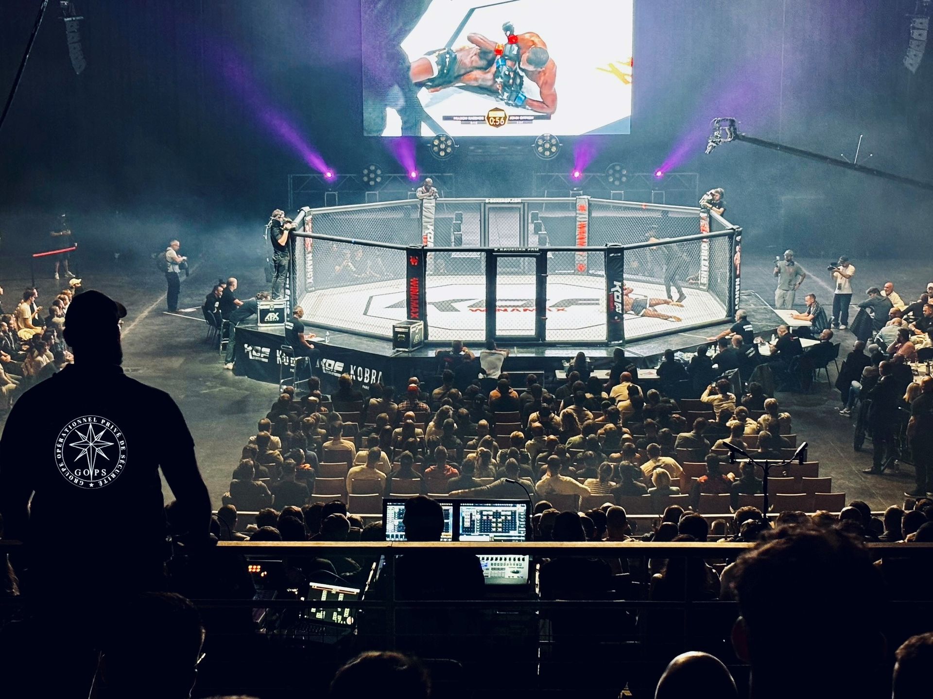 Combat de MMA dans une cage avec spectateurs, projecteurs et écran diffusant l'action.