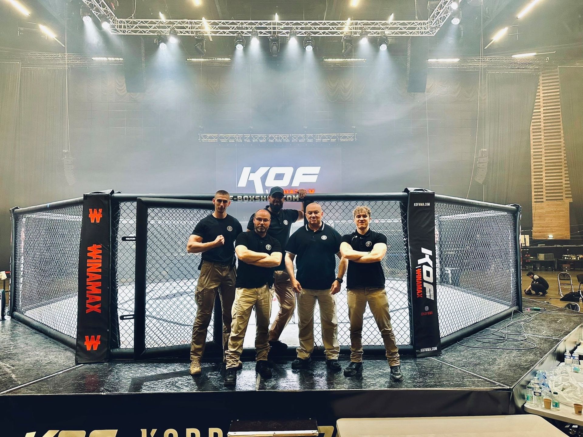 Des hommes en équipement tactique se tiennent devant une cage de MMA lors d'un événement, avec une scène en arrière-plan.