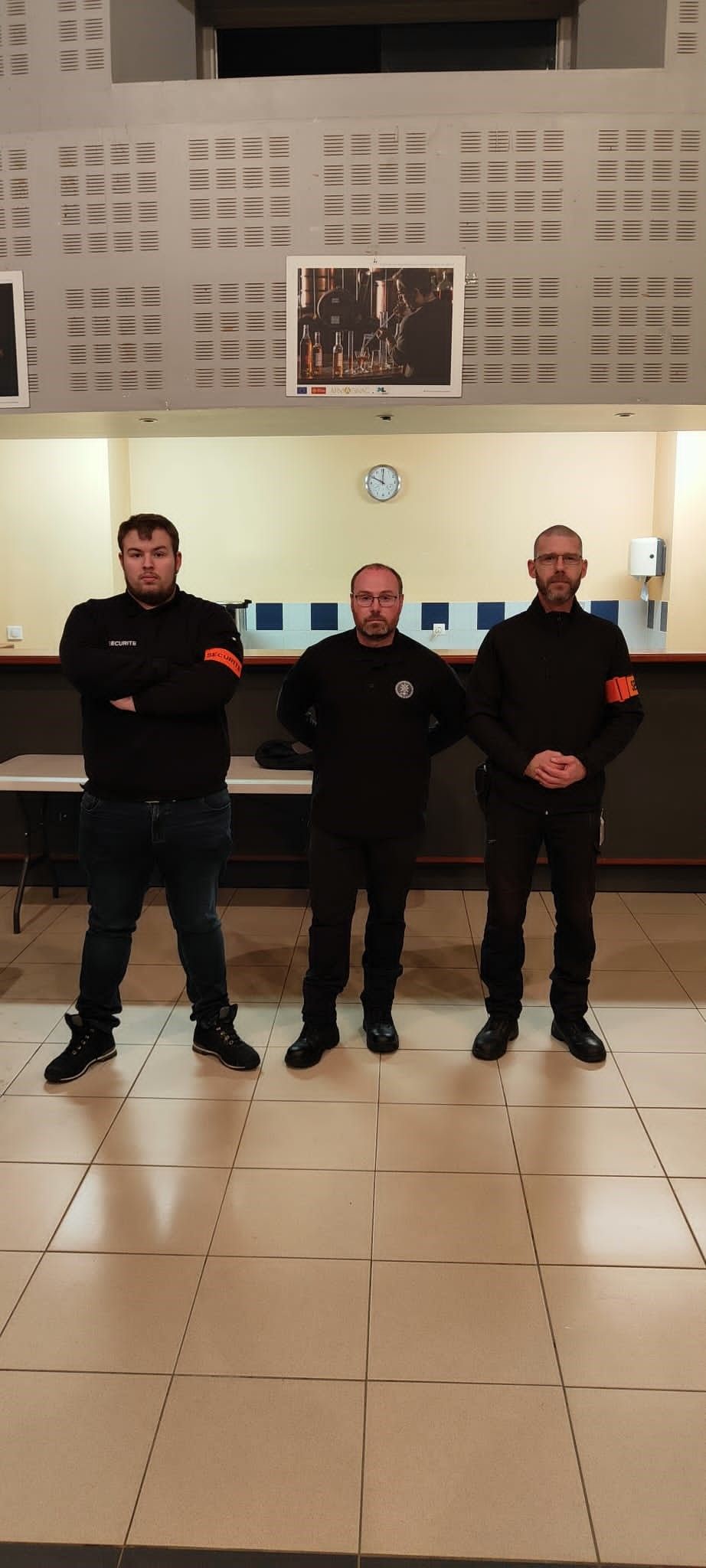 Trois hommes vêtus de noir se tiennent à l'intérieur. L'un a les bras croisés, les deux autres portent des brassards rouges.