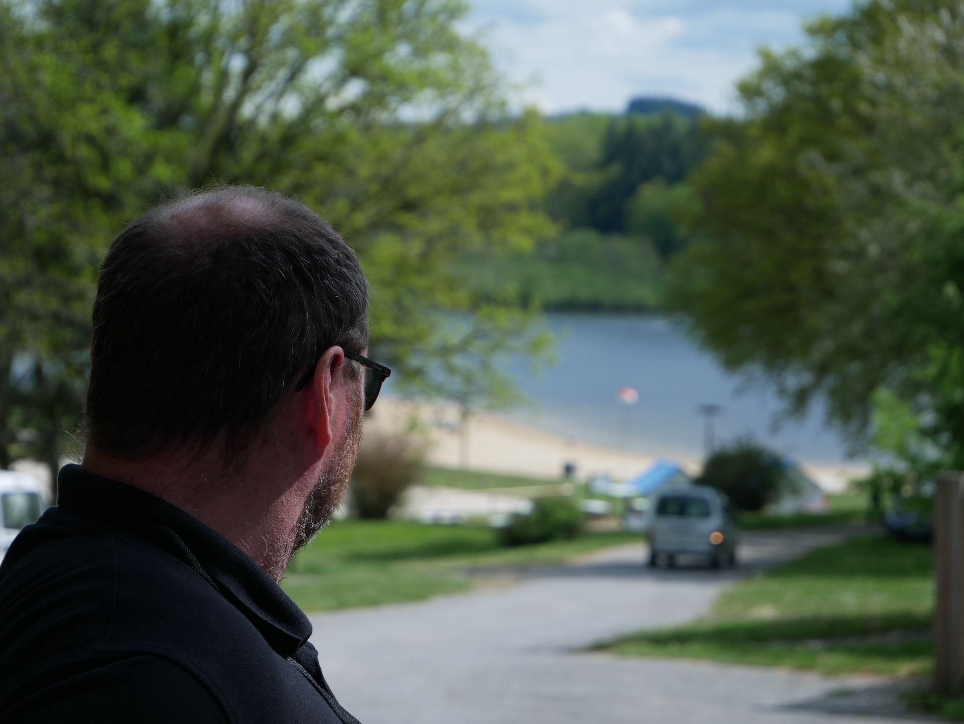 Homme portant des lunettes de soleil, regardant un lac et une route avec un véhicule. Arbres en arrière-plan.