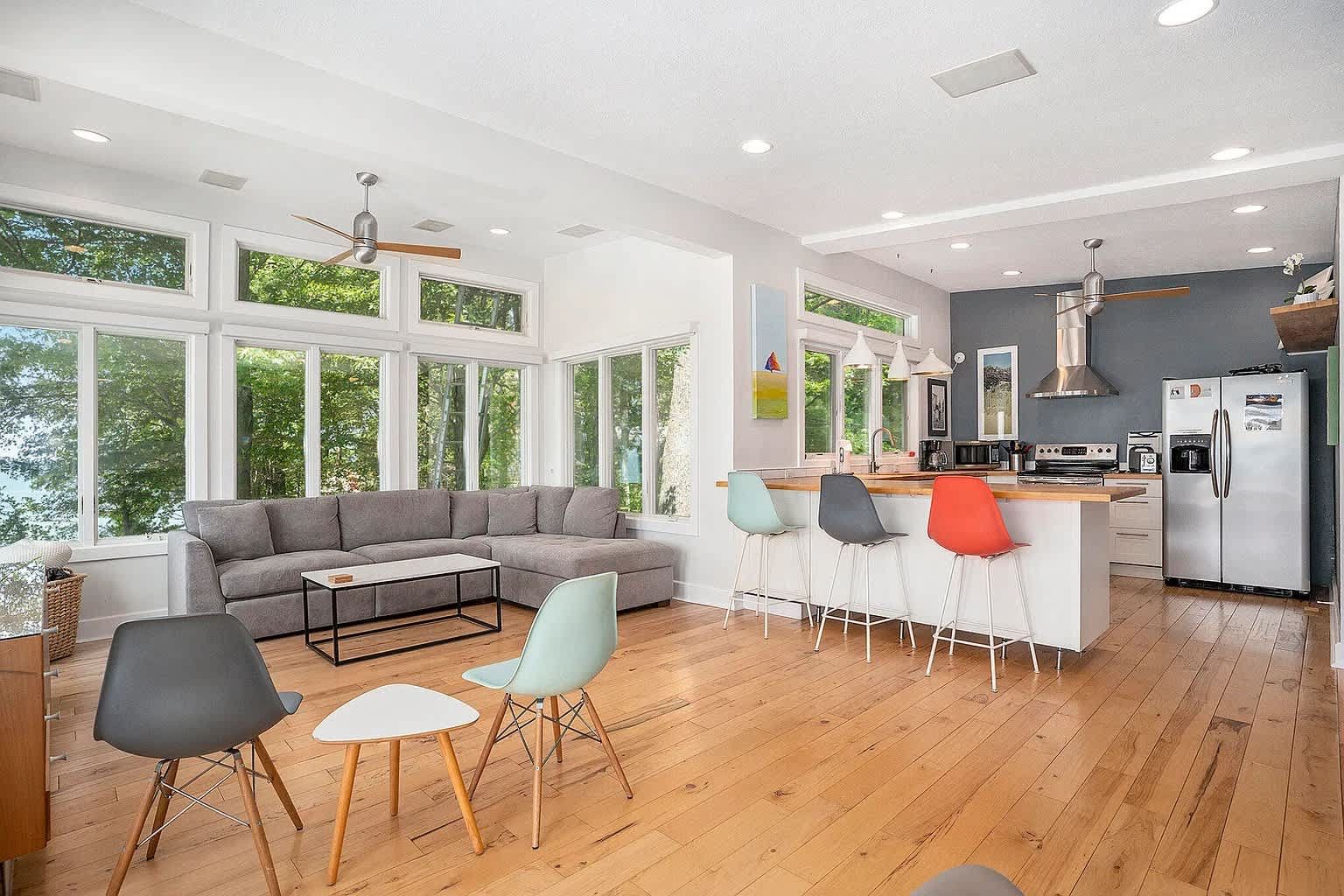 Spacious, bright living room with kitchen island, large windows, wood floors, and colorful chairs.