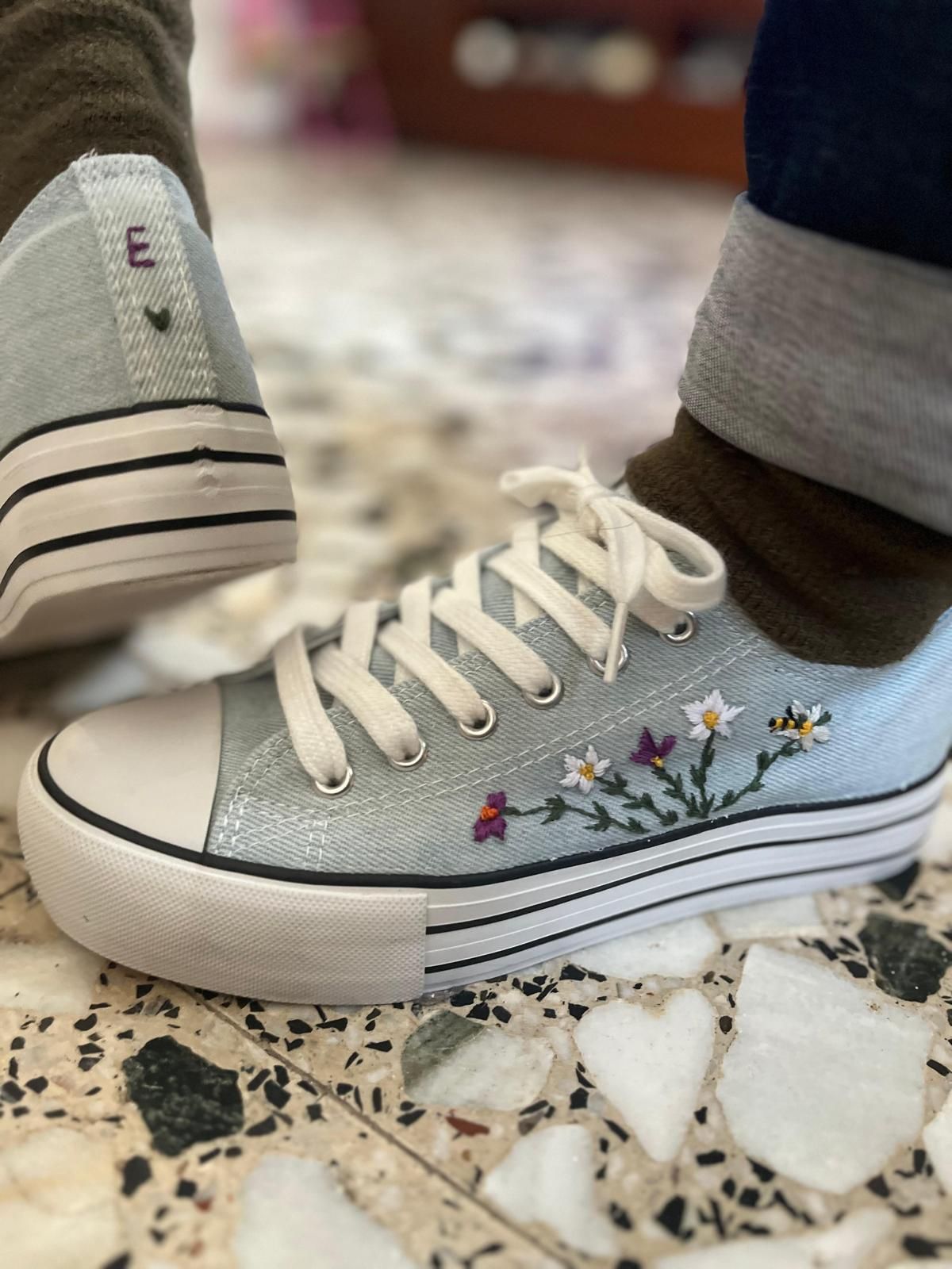 Zapatillas blancas con purpurina y bordados florales, mostradas sobre un suelo de baldosas moteadas, con un pie que lleva una sola zapatilla.