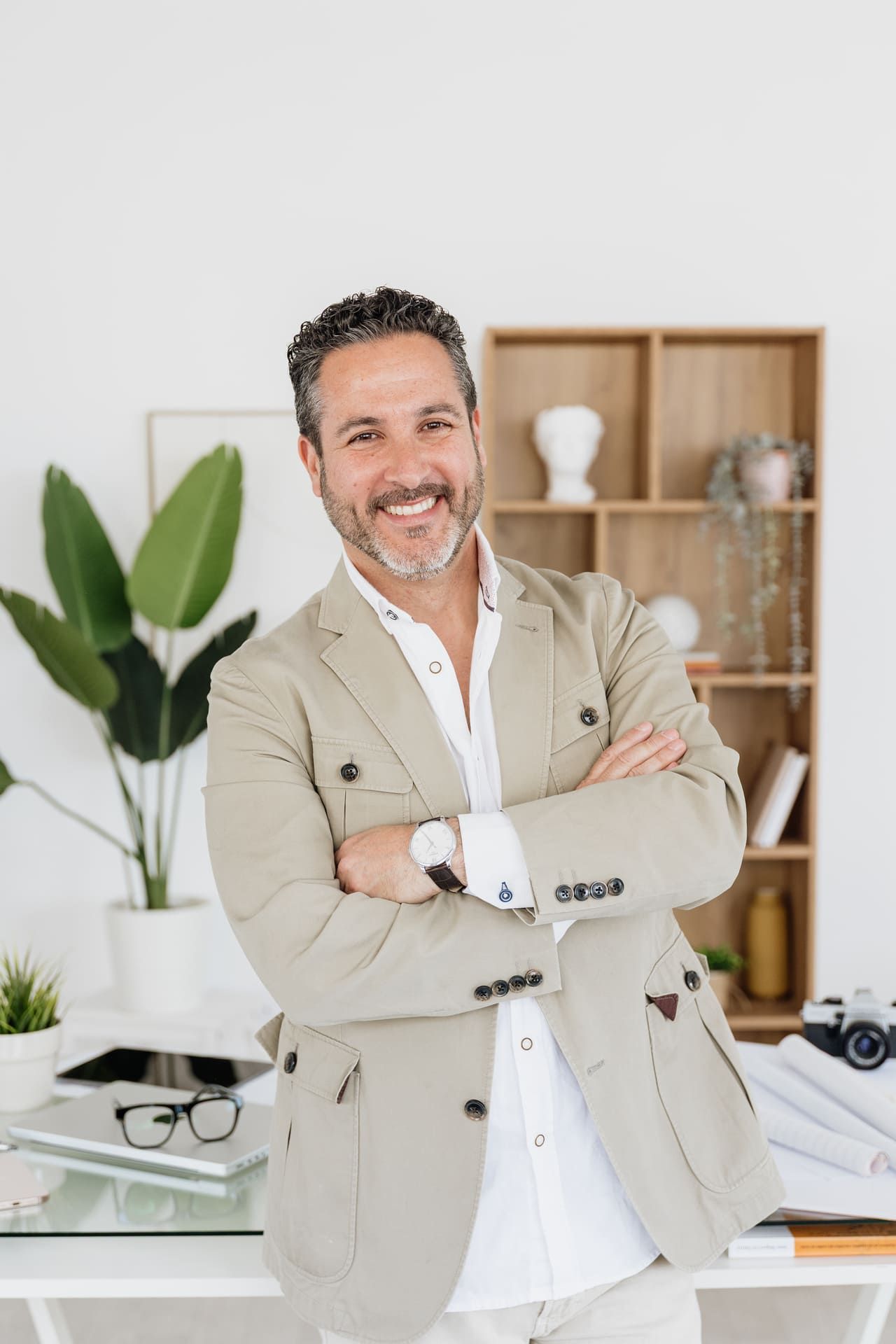 Hombre sonriente con chaqueta beige de pie con los brazos cruzados en una oficina luminosa.