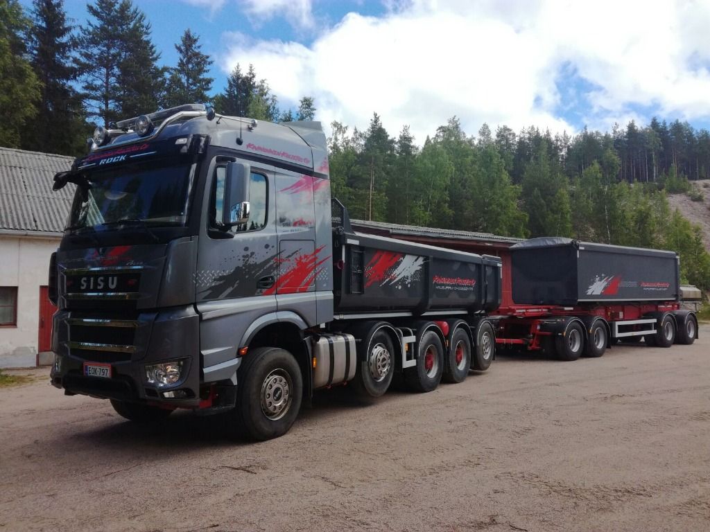 Harmaa Sisu-kuorma-auto vetää punaisilla yksityiskohdilla varustettua perävaunua pysäköitynä sorakentälle.