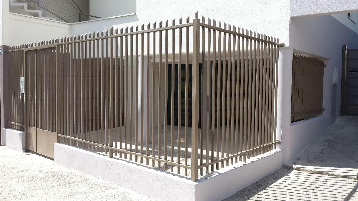 Une clôture métallique entoure l'angle d'un bâtiment blanc. La clôture est brune et une porte se trouve sur le côté gauche.