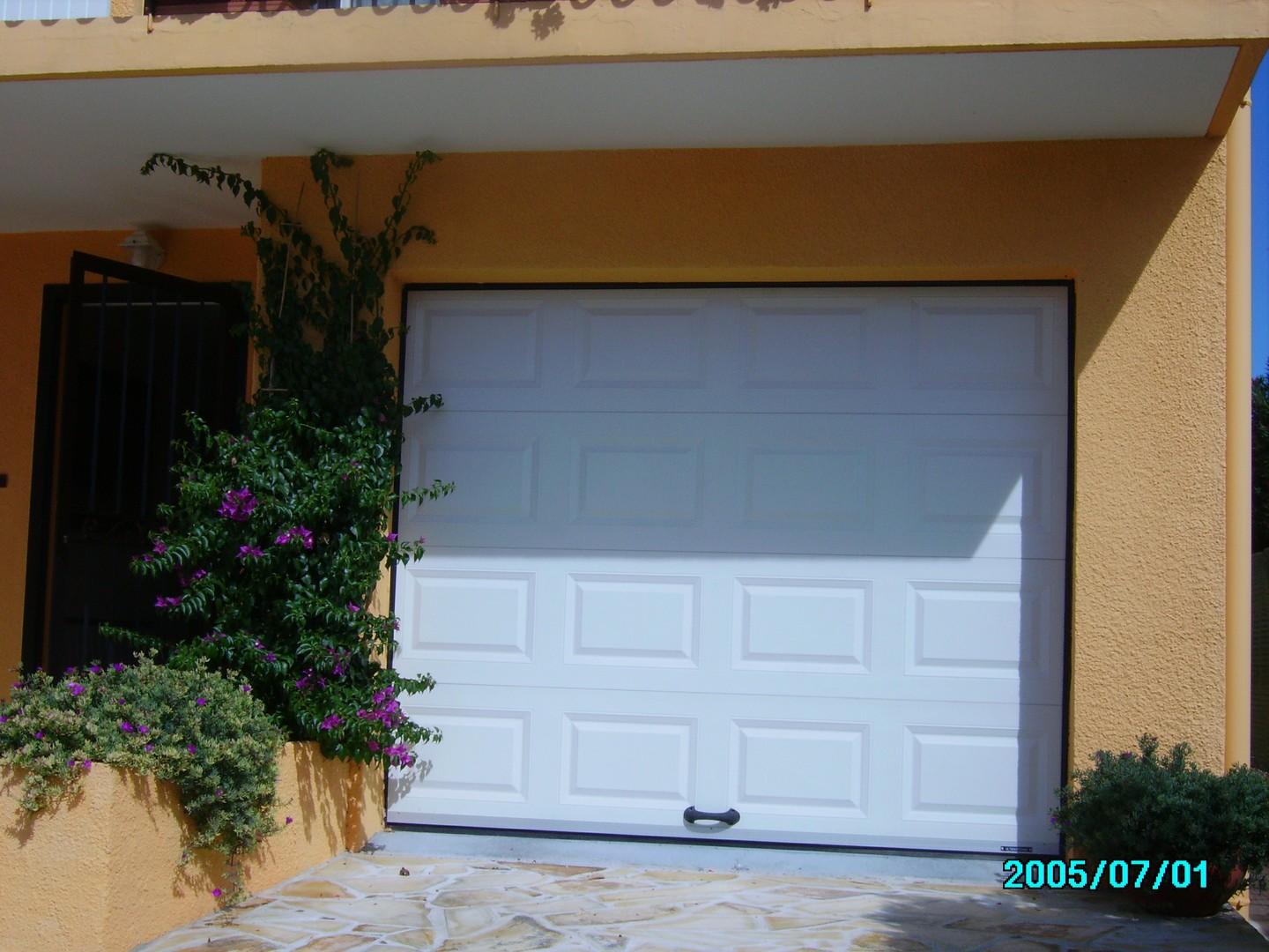 Porte de garage blanche sur un bâtiment jaune, avec des plantes vertes sur le côté.