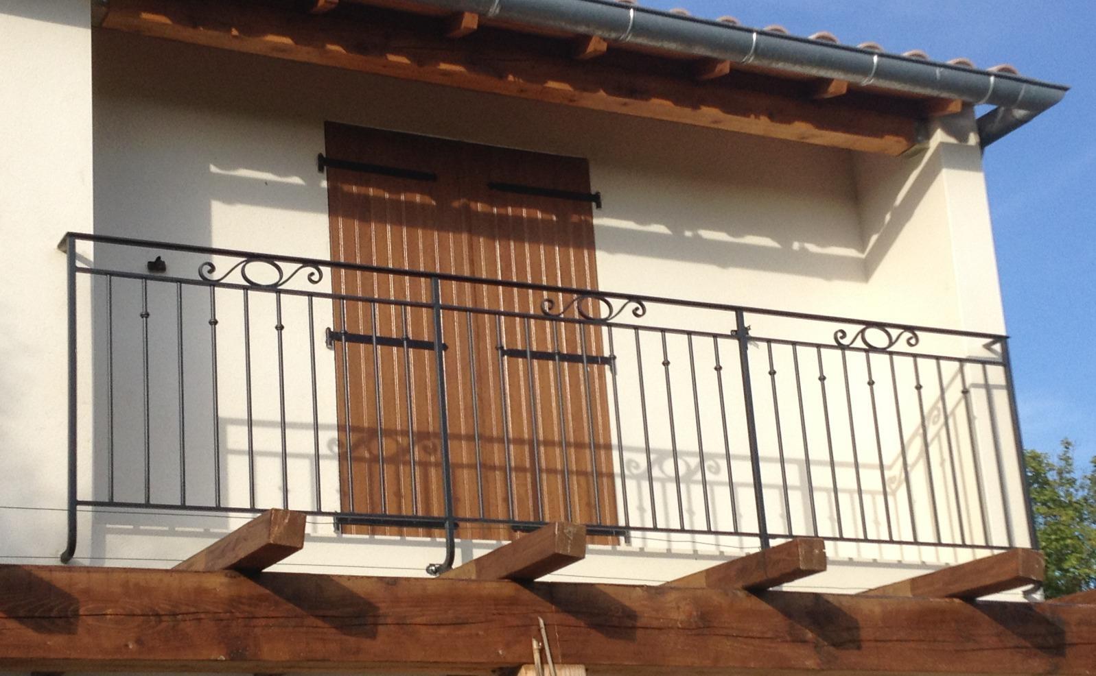 Rambarde de balcon en fer forgé noir sur une maison blanche aux volets marron.