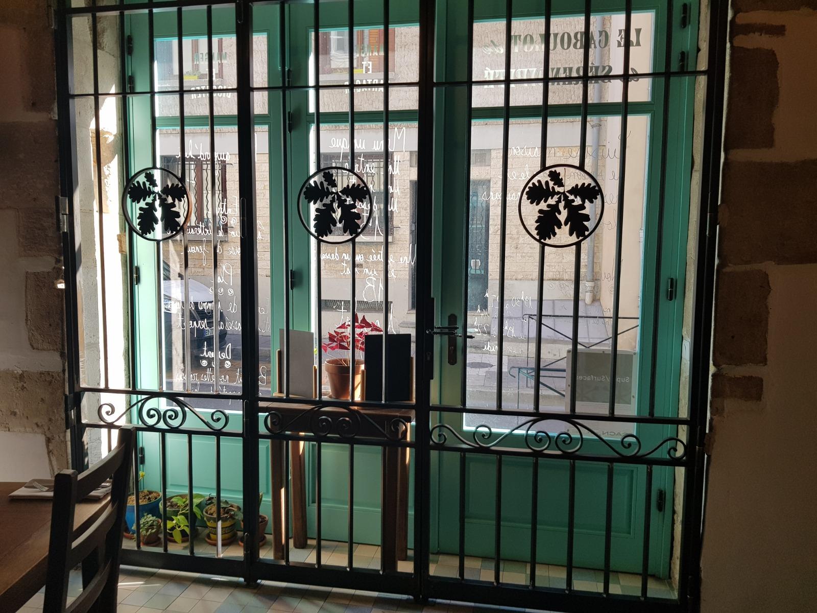 Double porte peinte en vert derrière des barreaux de sécurité en métal noir. Une petite table, sur laquelle reposent des livres et des fleurs, est visible à travers les portes.