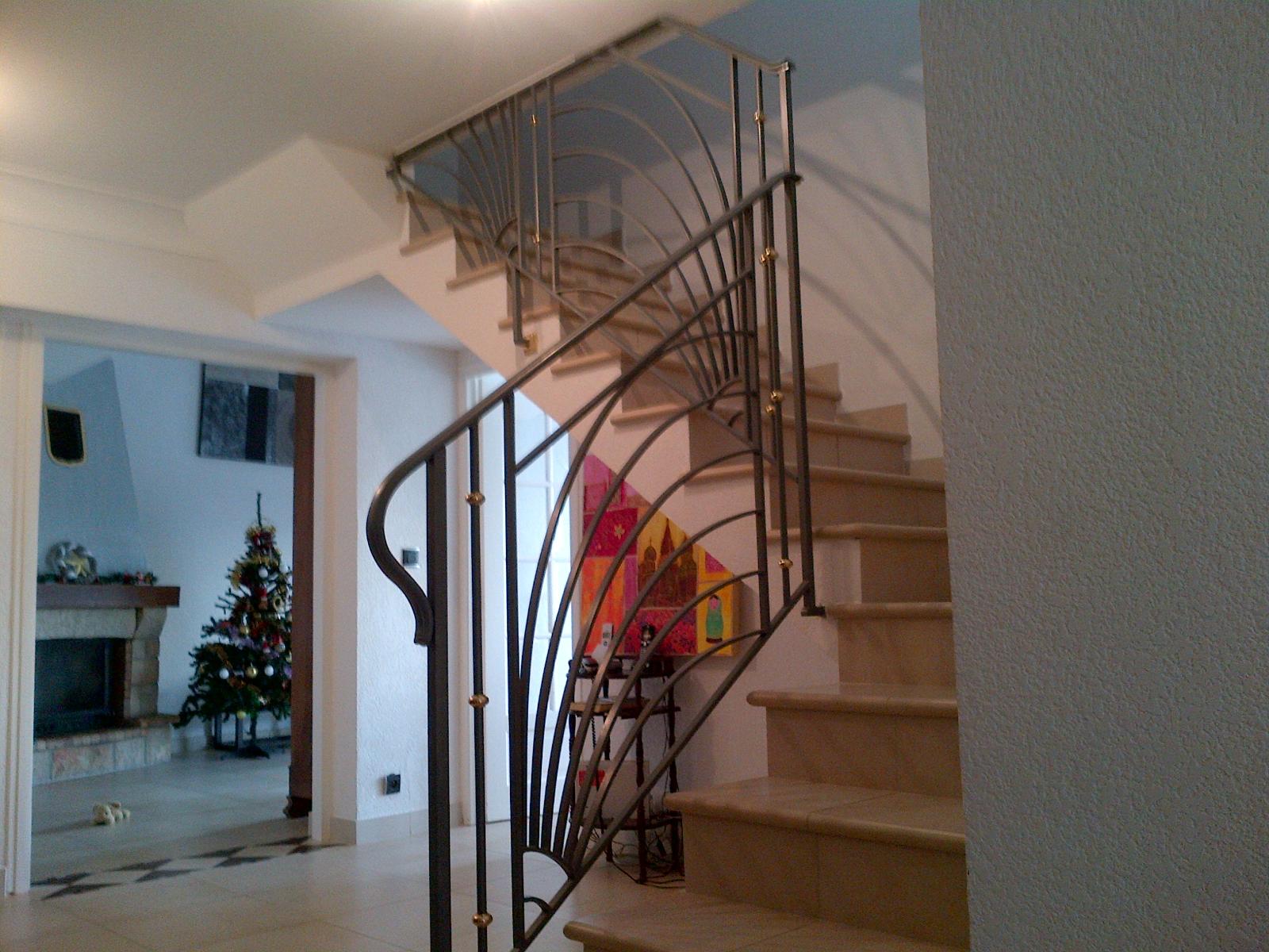 Escalier avec rampe en fer forgé ouvragée. Marches en marbre beige, murs blancs et un aperçu d'un sapin de Noël.