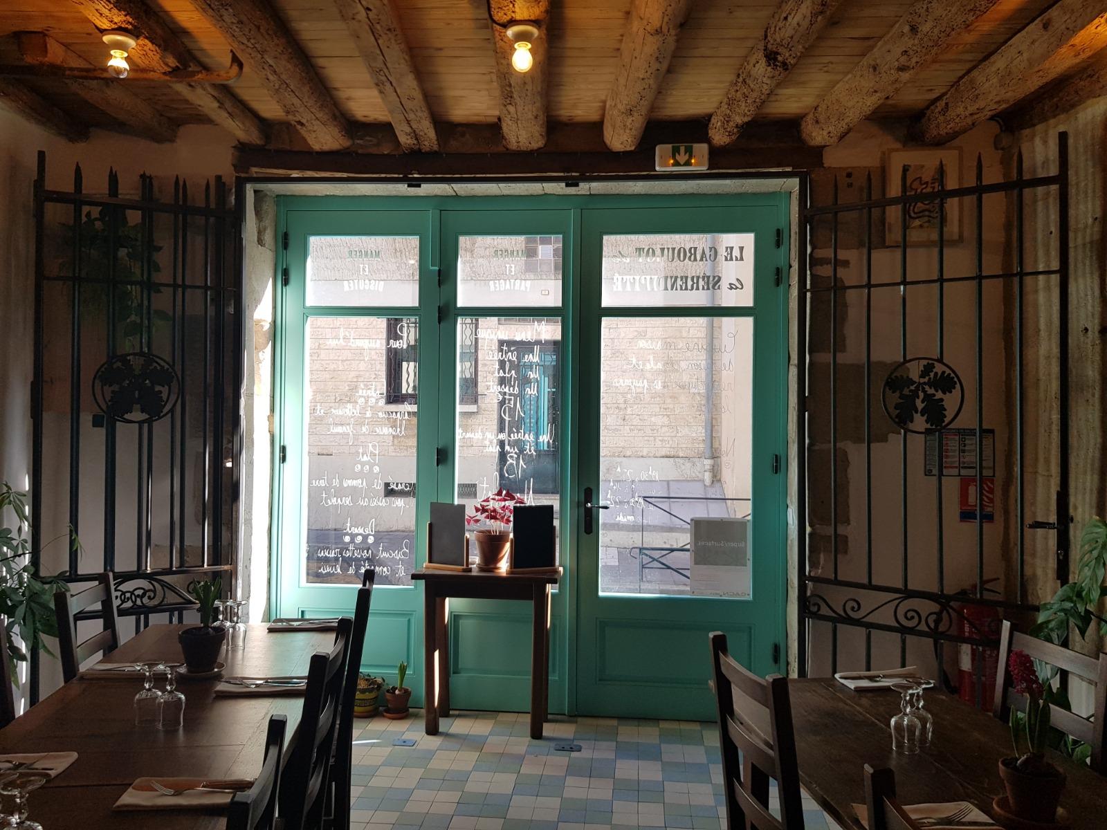 Intérieur du restaurant avec une porte turquoise donnant sur l'extérieur, des tables en bois et des poutres apparentes au plafond.