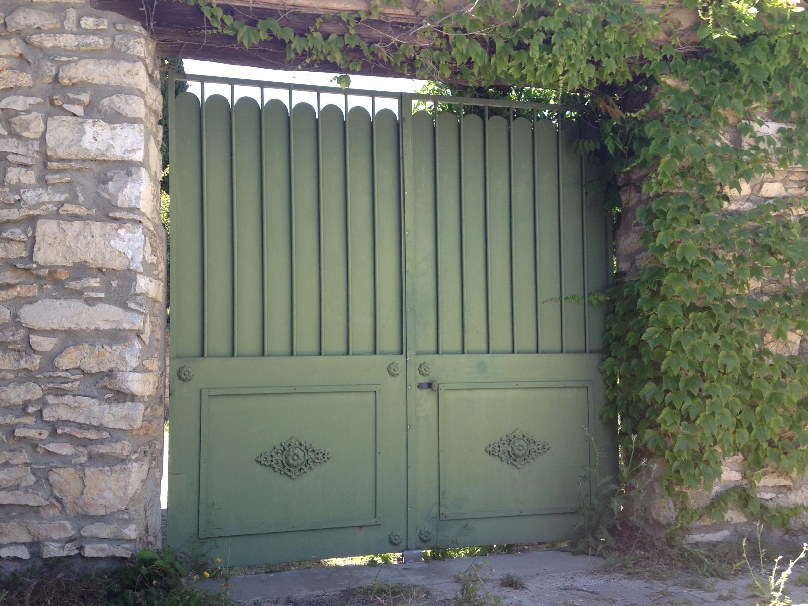 Portail en métal vert encadré par des murs de pierre, avec des vignes qui poussent sur la droite.