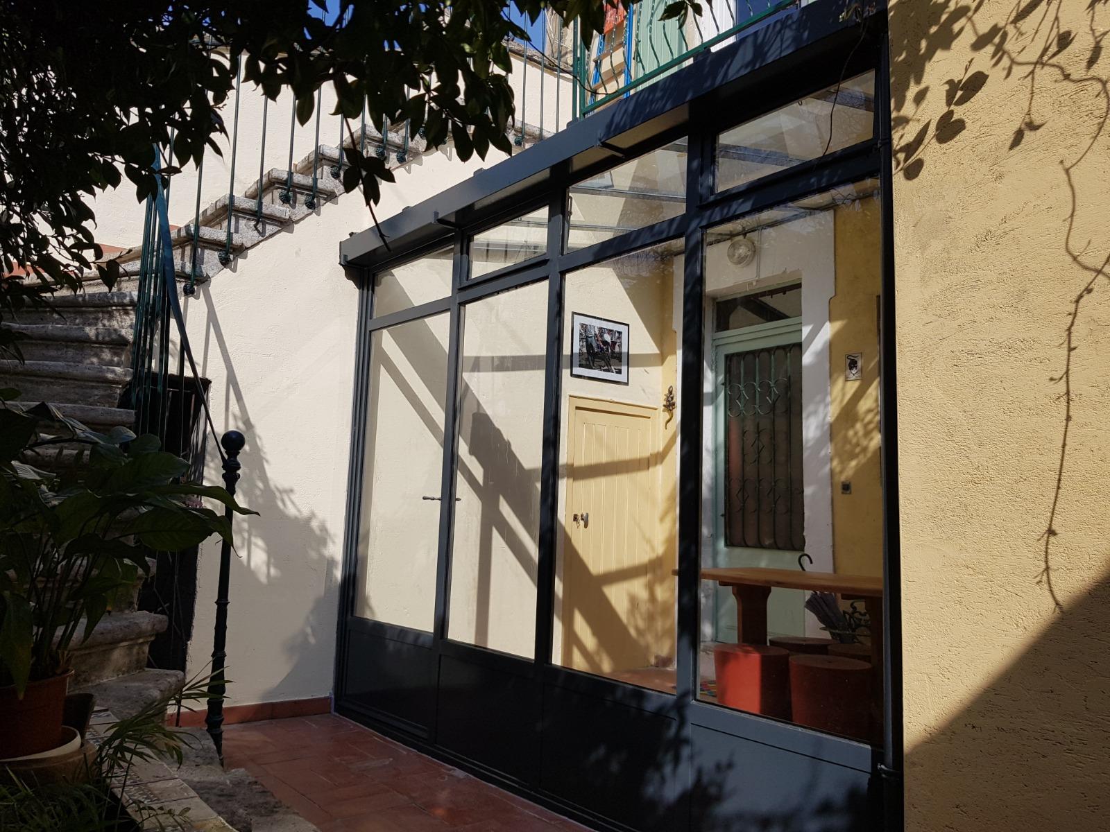 Une véranda avec une structure noire, des panneaux de verre et une vue sur une table et une chaise à l'intérieur. Mur jaune.