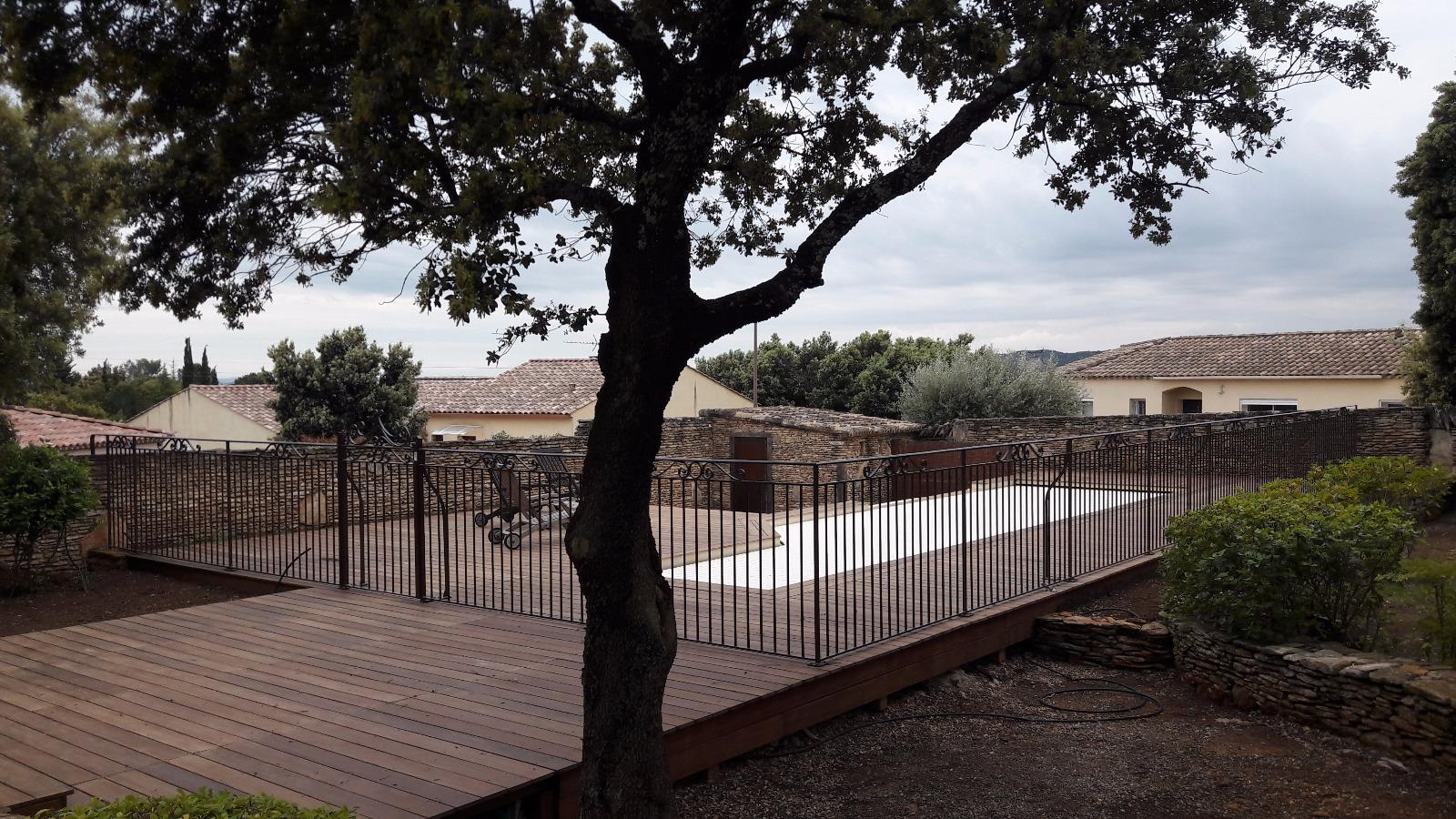 Une terrasse en bois et une clôture entourent une piscine, avec des bâtiments en arrière-plan sous un ciel nuageux.