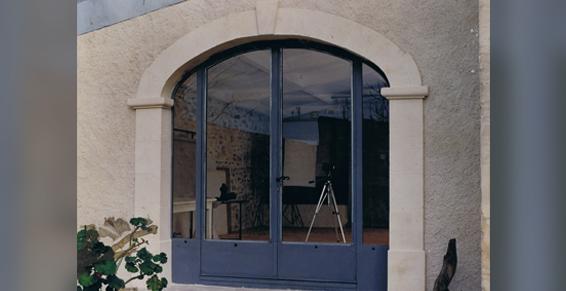 Porte cintrée avec portes vitrées à plusieurs panneaux, entourée d'encadrements en pierre et d'un mur extérieur en stuc.