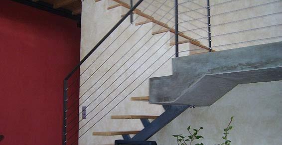 Escalier avec marches en bois et palier en béton ; rampe et support en métal noir. Mur rouge à gauche.