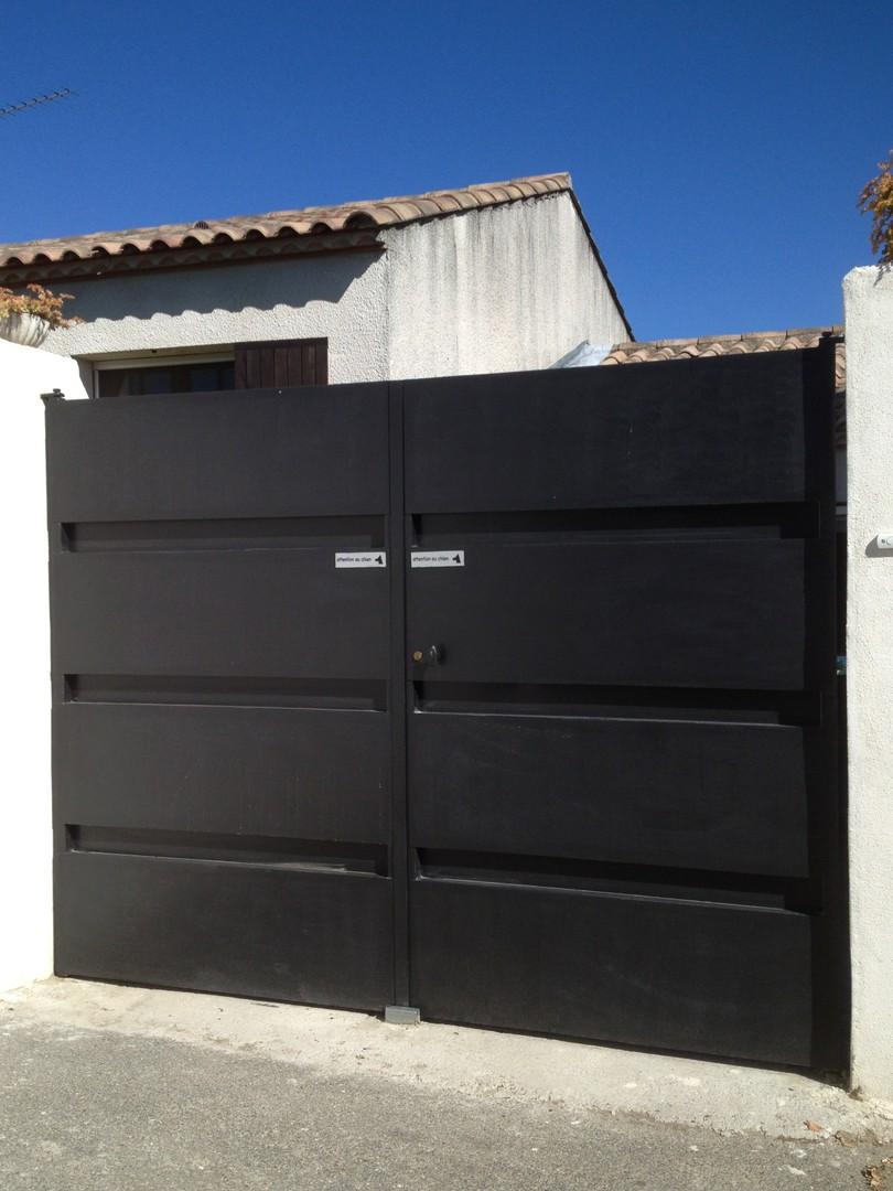 Portail en métal noir à barreaux horizontaux, se détachant sur un mur blanc et un bâtiment au toit de tuiles.
