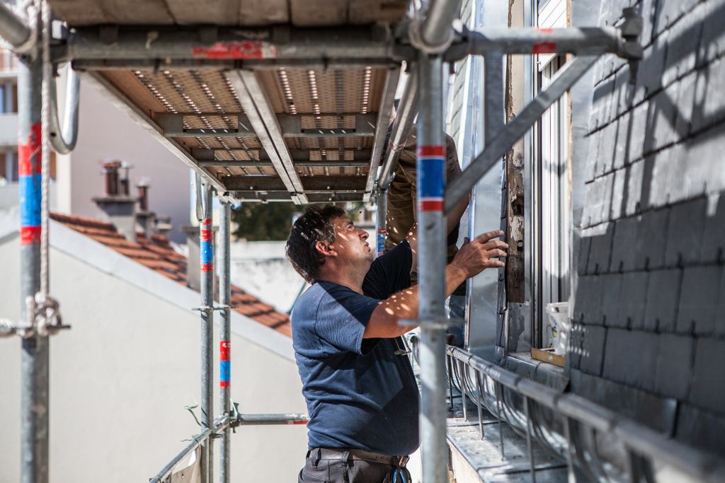 Ouvrier sur un échafaudage appliquant un enduit sur un mur de briques à côté d'un bâtiment blanc