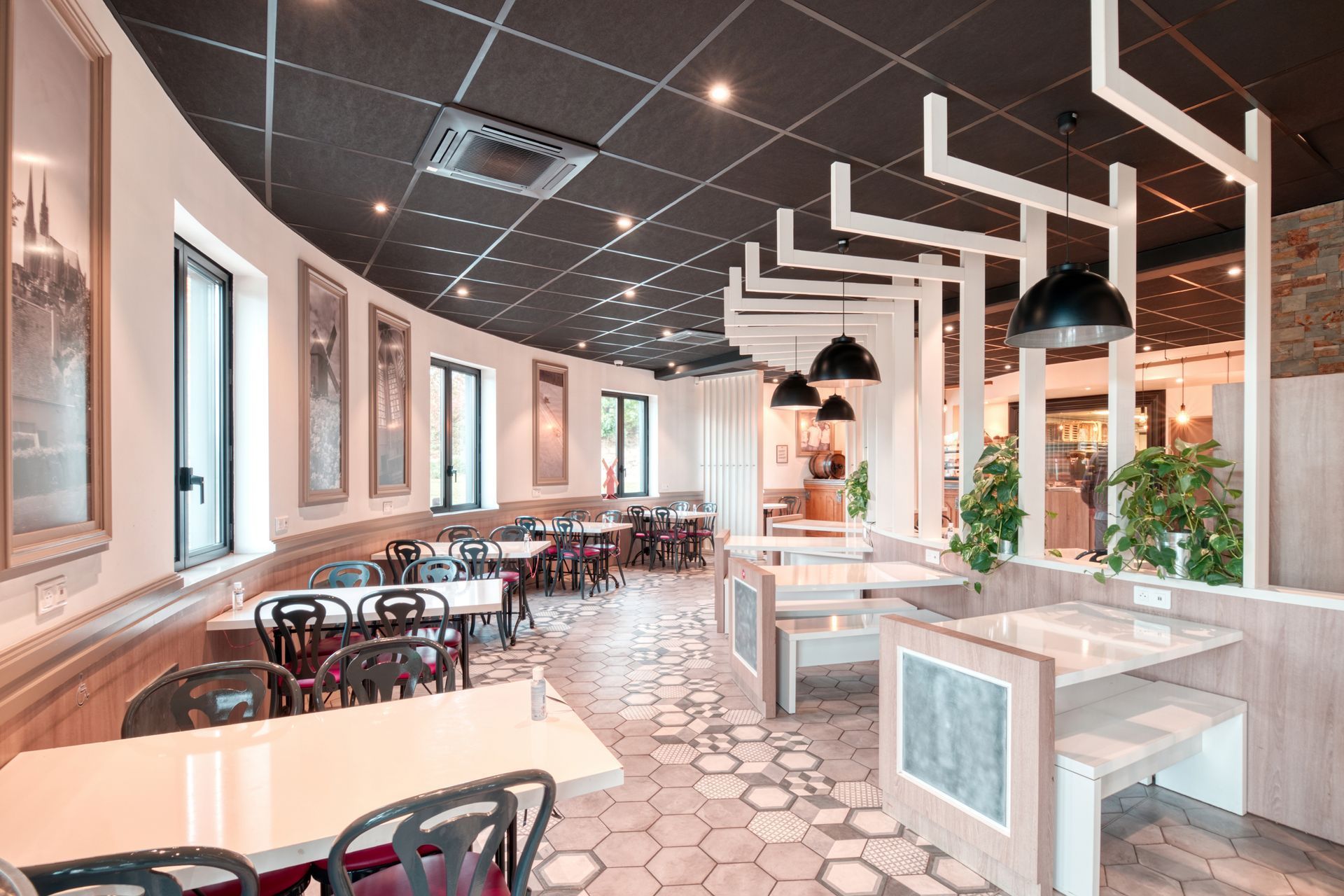 Intérieur de café moderne avec tables claires, plafond sombre et carrelage hexagonal au sol, aménagé avec des banquettes et des chaises.
