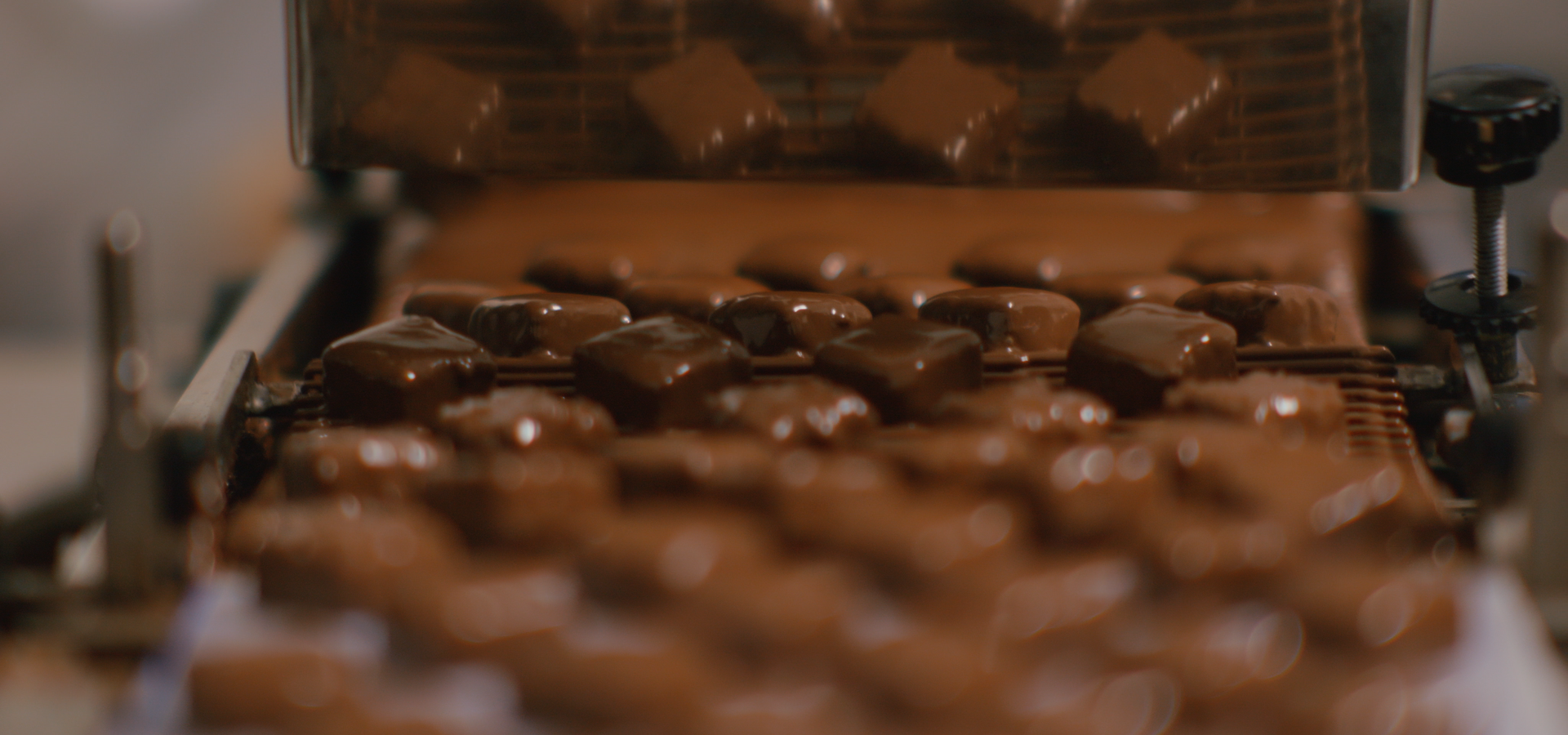 Dans une usine agroalimentaire, des rangées de carrés de chocolat défilent sur un tapis roulant, pressées par un outil métallique à motifs.