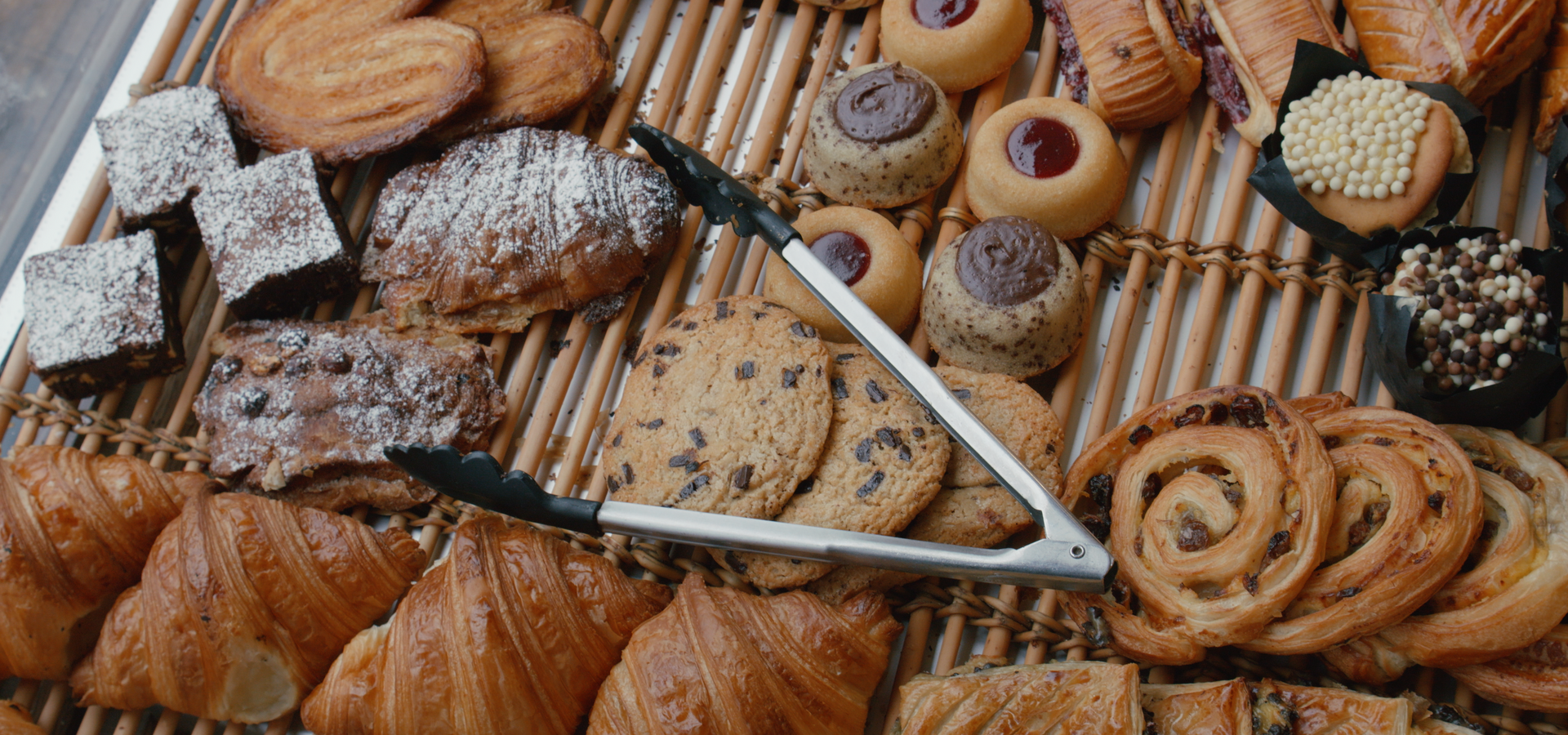 Un assortiment de pâtisseries, de biscuits et de brownies disposés sur un support en bois, avec des pinces en métal posées au centre.