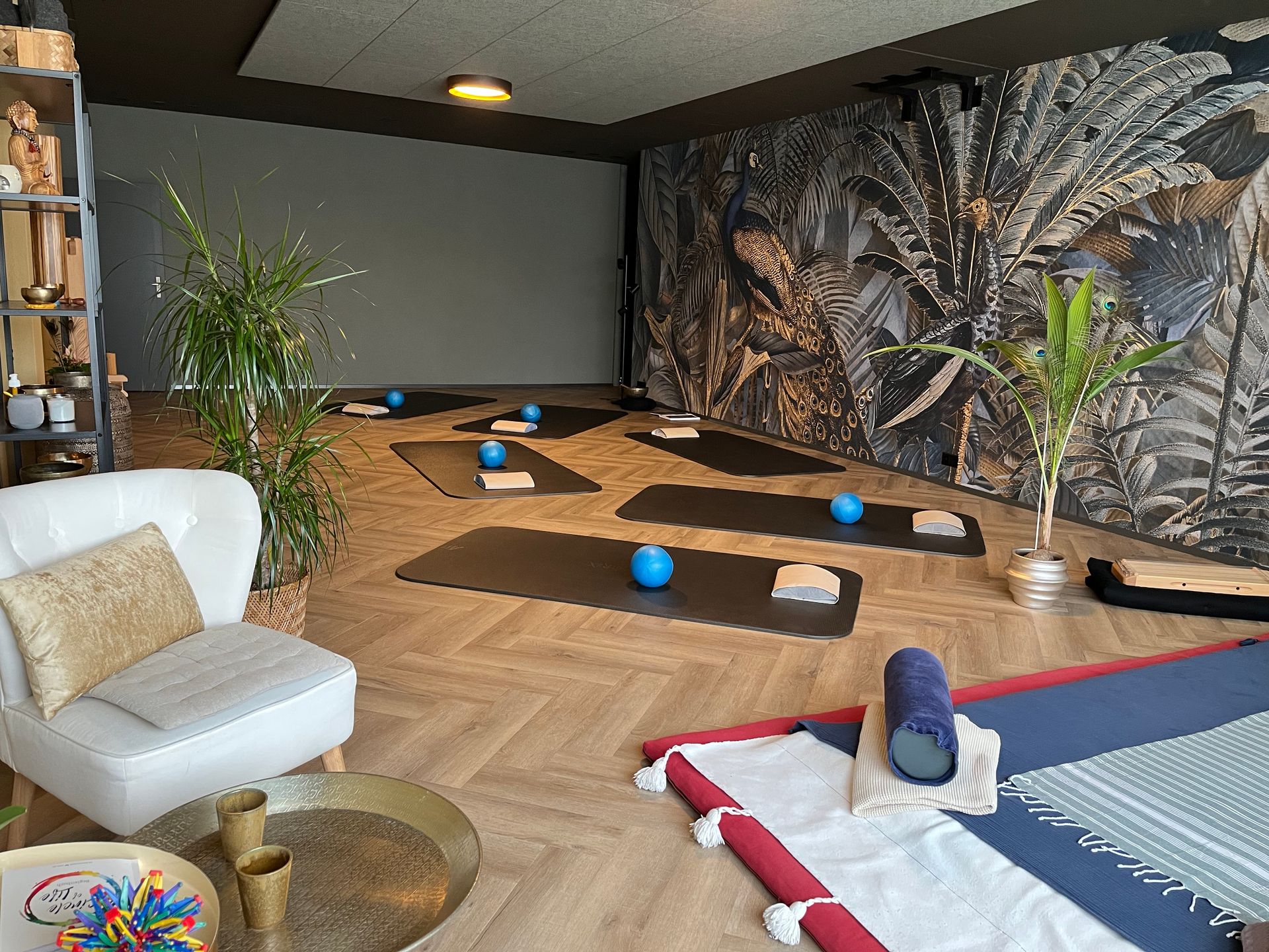 Yogastudio mit Matten, Requisiten, Pflanzen und einem weißen Stuhl; warme Beleuchtung und gemusterte Tapete.