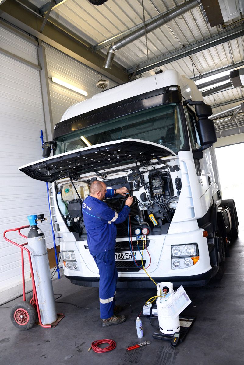Un mécanicien en combinaison bleue travaille sur un camion blanc dans un garage, capot ouvert