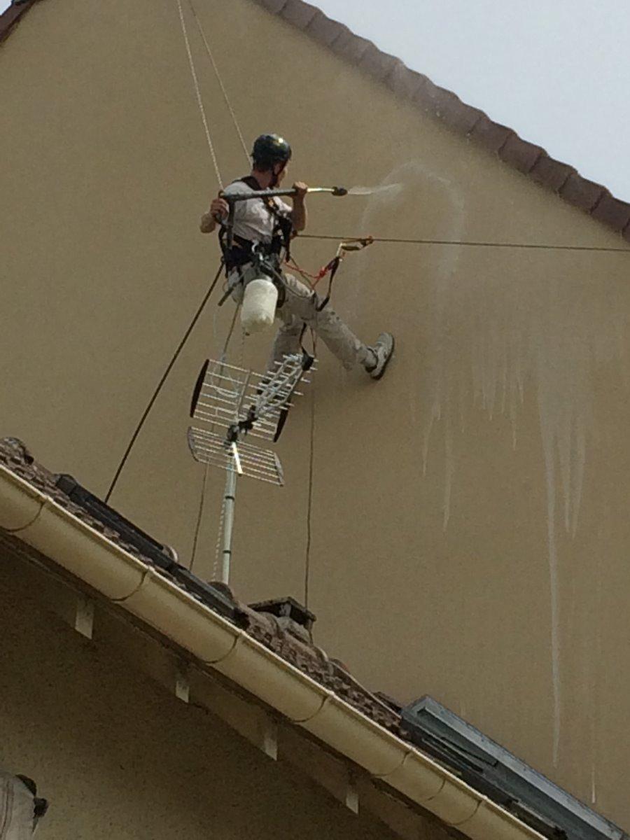 Une personne nettoie un mur extérieur à haute pression, suspendue par des cordes.