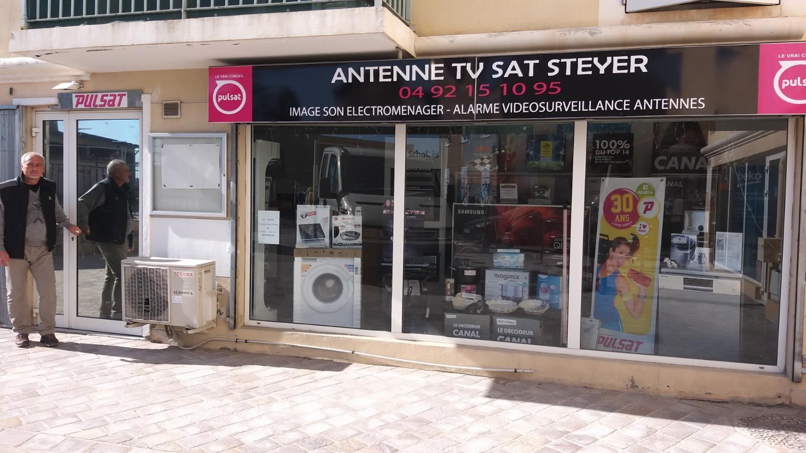 Devanture avec deux hommes debout à l'extérieur, panneau indiquant « ANTENNE TV SAT STEYER ». Présentoirs de téléviseurs, d'appareils électroménagers et d'antennes.