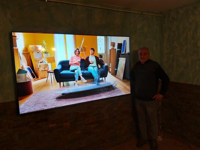 Un homme se tient à côté d'un grand téléviseur montrant un couple sur un canapé dans une pièce avec des cartons de déménagement.