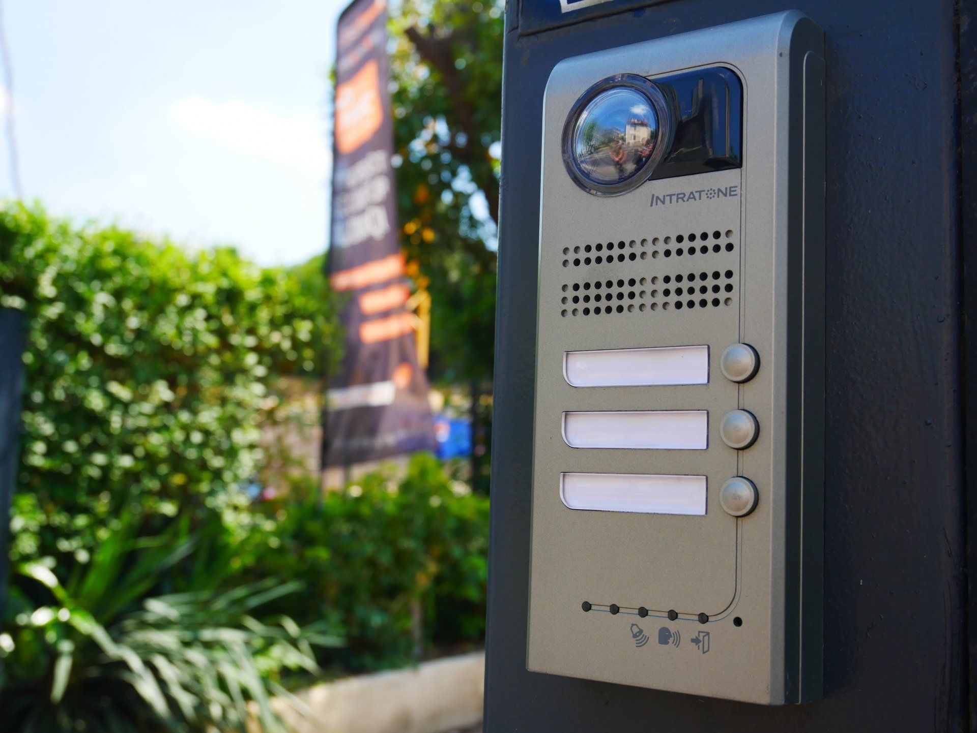 Interphone extérieur avec caméra, haut-parleur et trois boutons, monté sur un mur près de la verdure