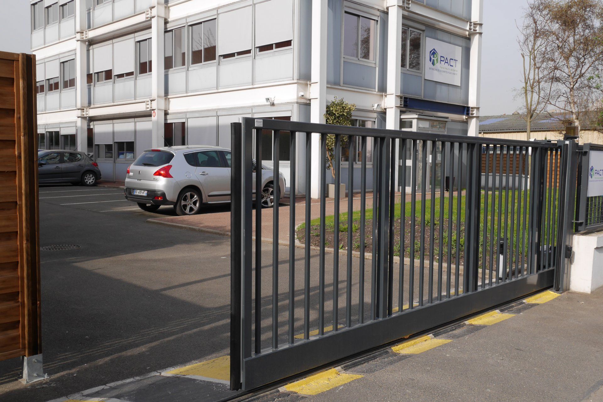 Portail coulissant en métal noir à l'entrée d'un immeuble, avec des voitures garées en arrière-plan