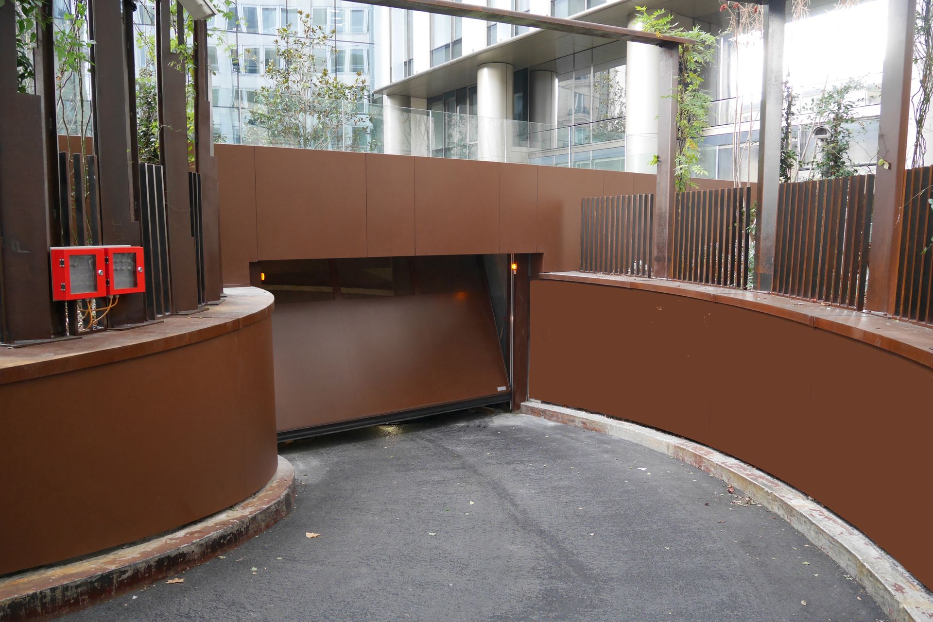 Rampe incurvée marron menant à l'entrée d'un parking souterrain avec des bornes d'appel d'urgence rouges